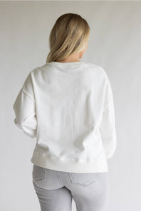 A woman viewed from behind wearing a plain white crewneck sweatshirt and light gray jeans, standing against a neutral light gray studio background.