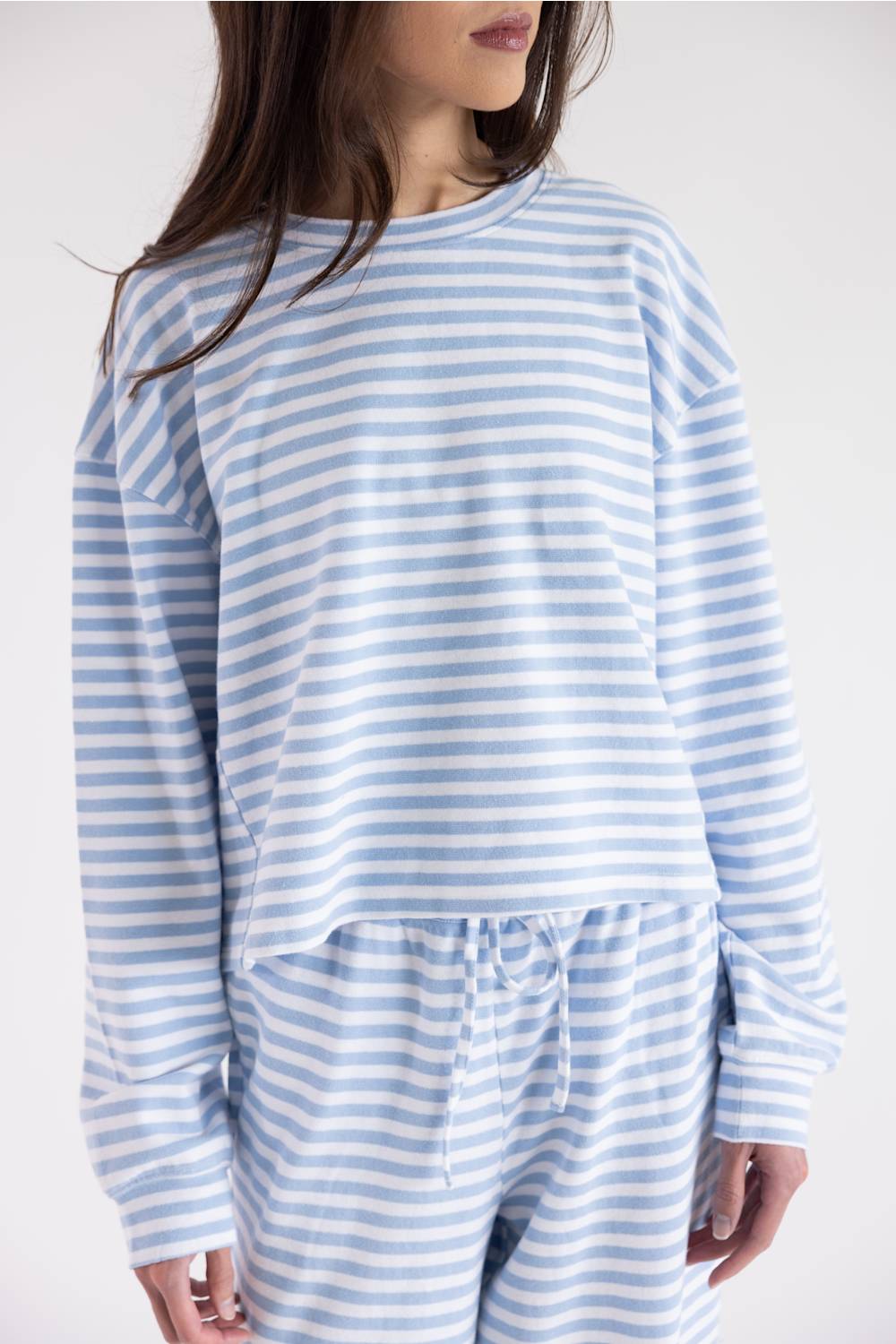 A woman wearing a light blue and white striped long-sleeve crop top and matching striped drawstring pants against a plain light gray studio background.