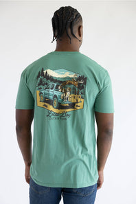 A photo taken from the back of a man in a light green shirt with a graphic of a truck with dogs, a deer carcass, and a cooler in the bed.