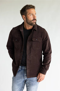 A man wearing a dark brown corduroy button-up shirt jacket over a black t-shirt and blue jeans, standing against a light gray studio background.