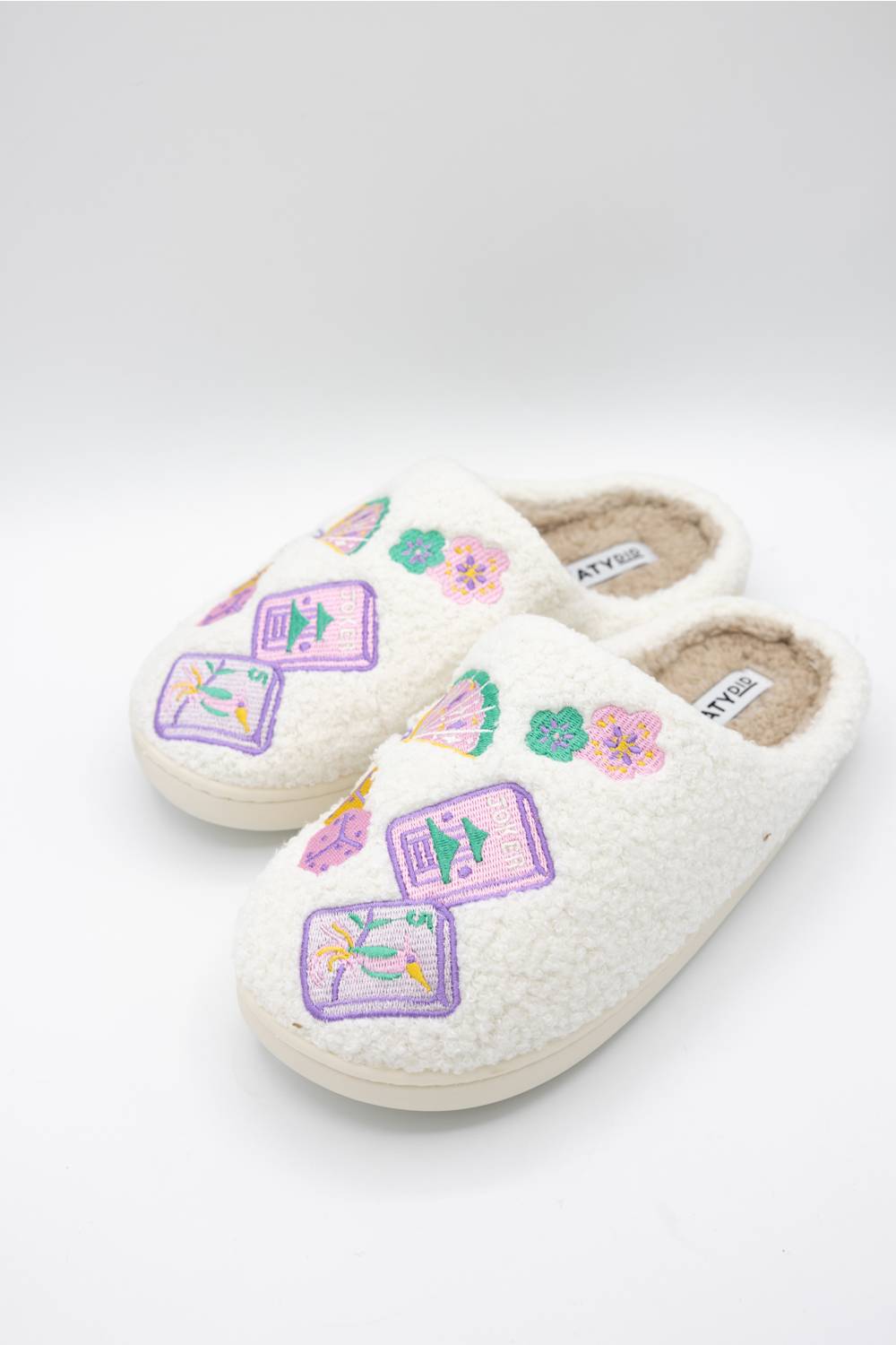 A pair of off-white sherpa slip-on slippers decorated with colorful majong and fans, displayed on a light gray background.