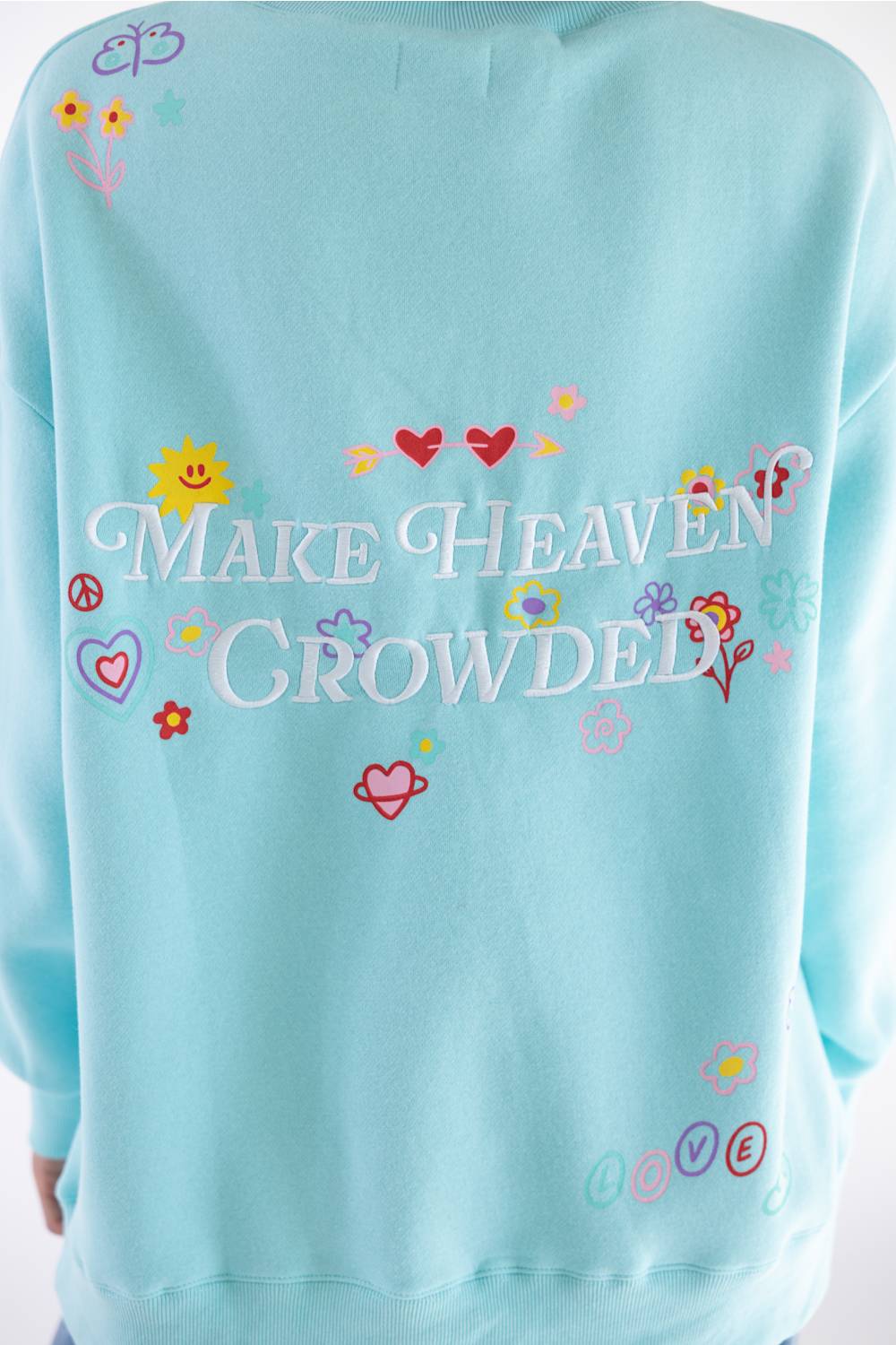 A light blue sweatshirt featuring the phrase 'MAKE HEAVEN CROWDED' in white script across the chest, surrounded by colorful embroidered hearts, flowers, and peace signs.