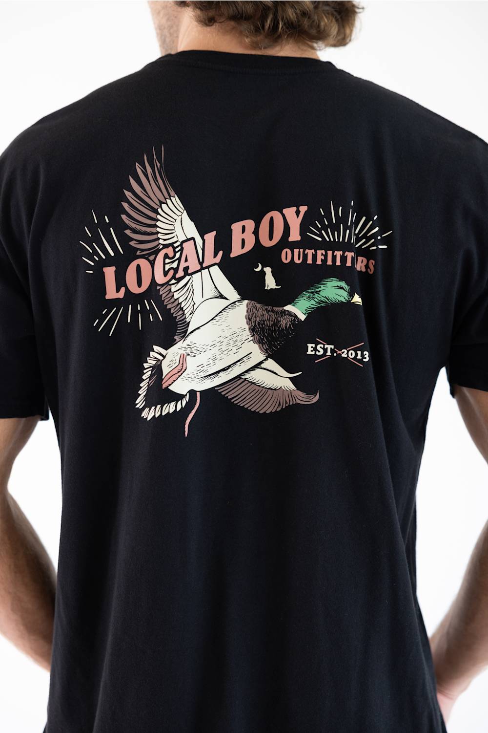 A man wearing a black t-shirt, viewed from the back. The shirt features a large graphic print of a flying mallard duck with the text 'LOCAL BOY OUTFITTERS' and 'EST. 2013