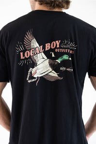 A man wearing a black t-shirt, viewed from the back. The shirt features a large graphic print of a flying mallard duck with the text 'LOCAL BOY OUTFITTERS' and 'EST. 2013
