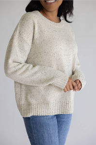 A woman wearing a cream-colored, speckled knit crewneck sweater with blue jeans, shown from the chest down against a plain light gray background.