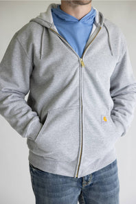 A man wearing a light gray zip-up hoodie with a drawstring hood, kangaroo pockets, and a small yellow Carhartt logo patch on the lower right front pocket. He is also wearing a light blue t-shirt and blue jeans, standing against a plain light gray wall.