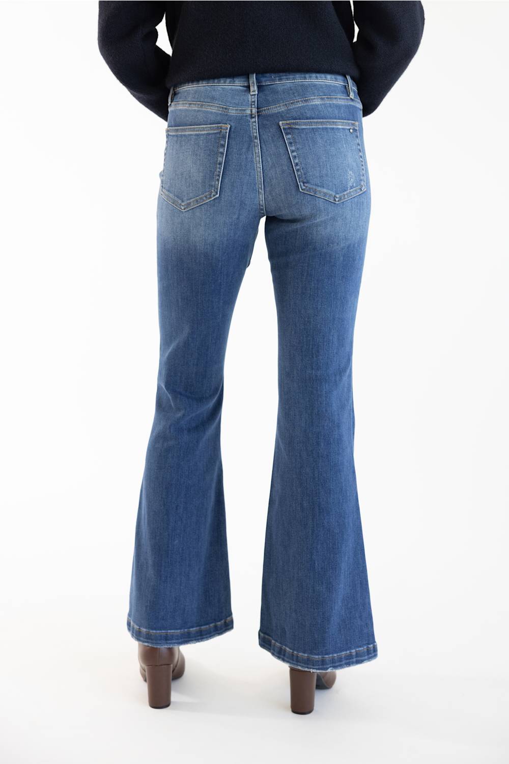 Rear view of a person wearing medium-dark blue flare jeans with a black top and brown heels, standing against a plain white background.