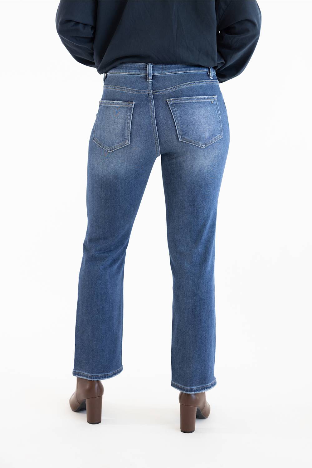 Rear view of a person wearing dark-wash blue jeans with a dark navy top and brown block-heel boots against a plain white studio background.