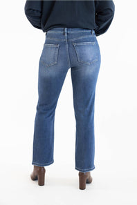 Rear view of a person wearing dark-wash blue jeans with a dark navy top and brown block-heel boots against a plain white studio background.