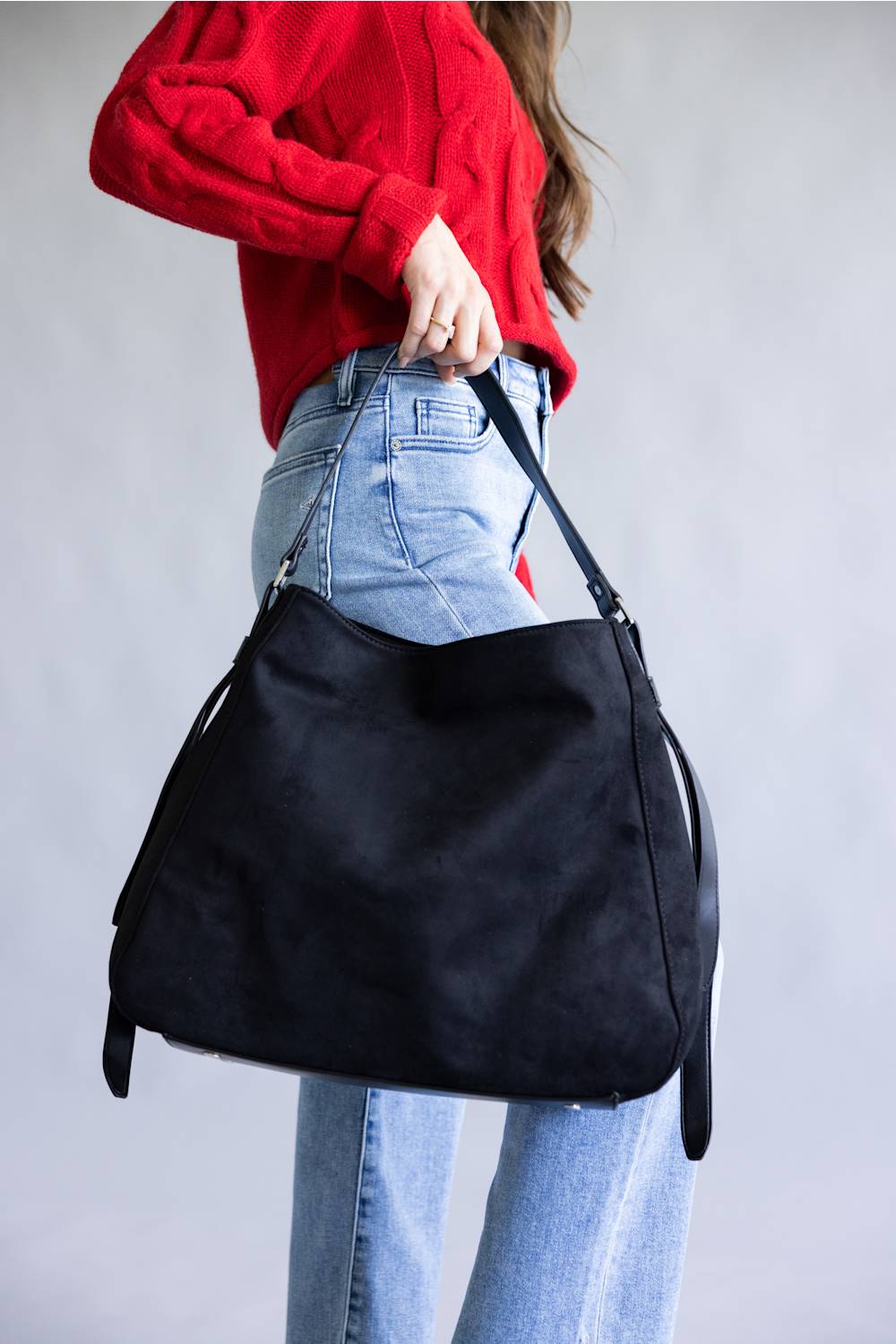 A person wearing a red knit sweater and light blue jeans carries a large black suede hobo shoulder bag against a plain gray studio background.