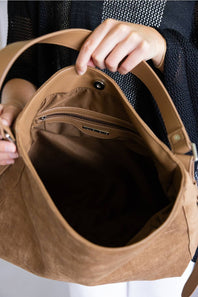 A close-up of a tan suede shoulder bag being held open by hands, showing a dark brown interior with a zippered pocket and snap closure.