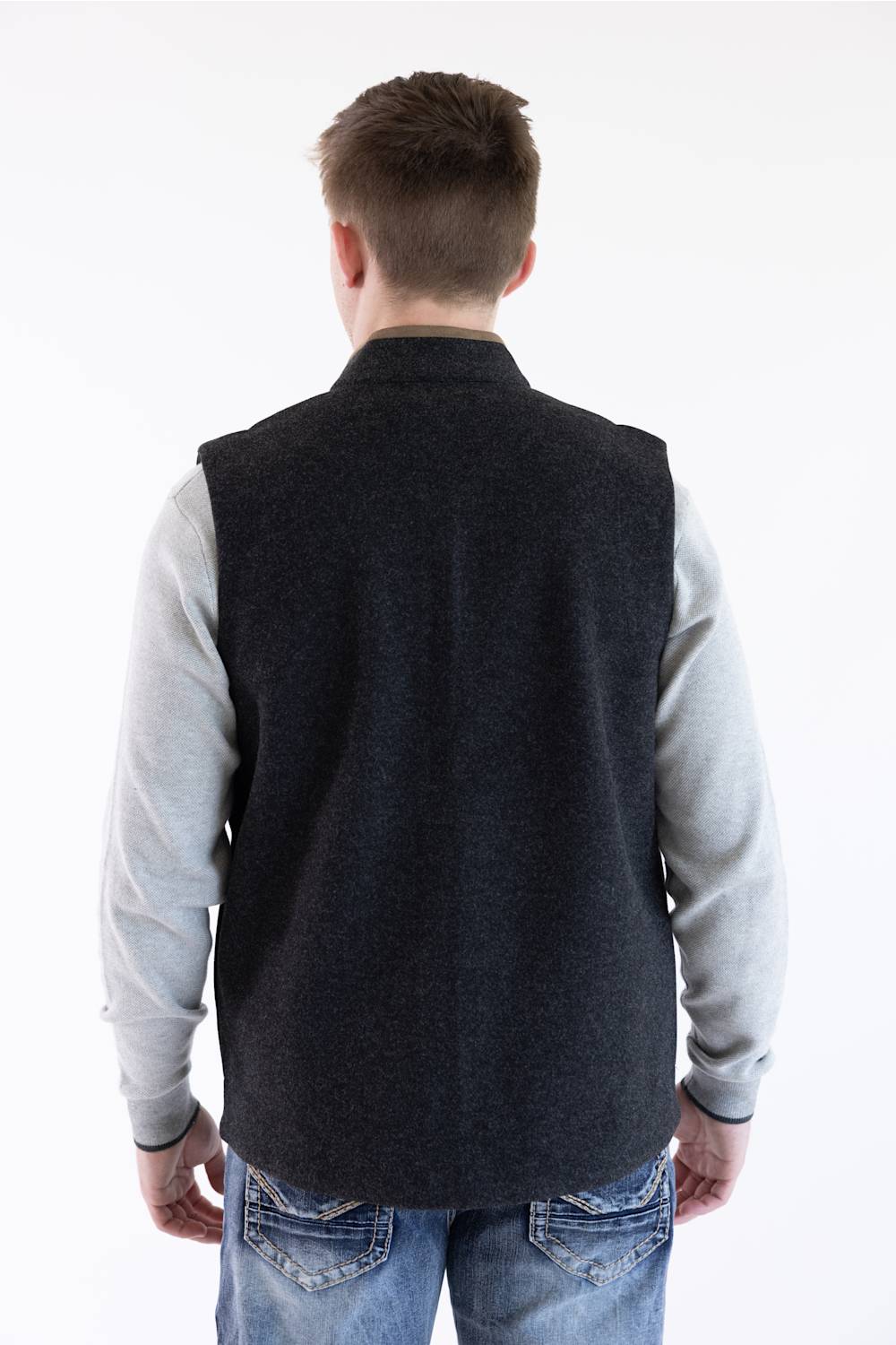 A man viewed from behind wearing a dark charcoal gray knit sweater vest over a light gray long-sleeve shirt and blue jeans, against a plain white background.