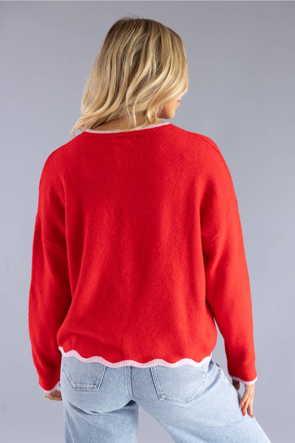 A woman viewed from behind wearing a bright red knit sweater with a scalloped white trim at the hem and cuffs, paired with light blue jeans, against a solid gray studio background.