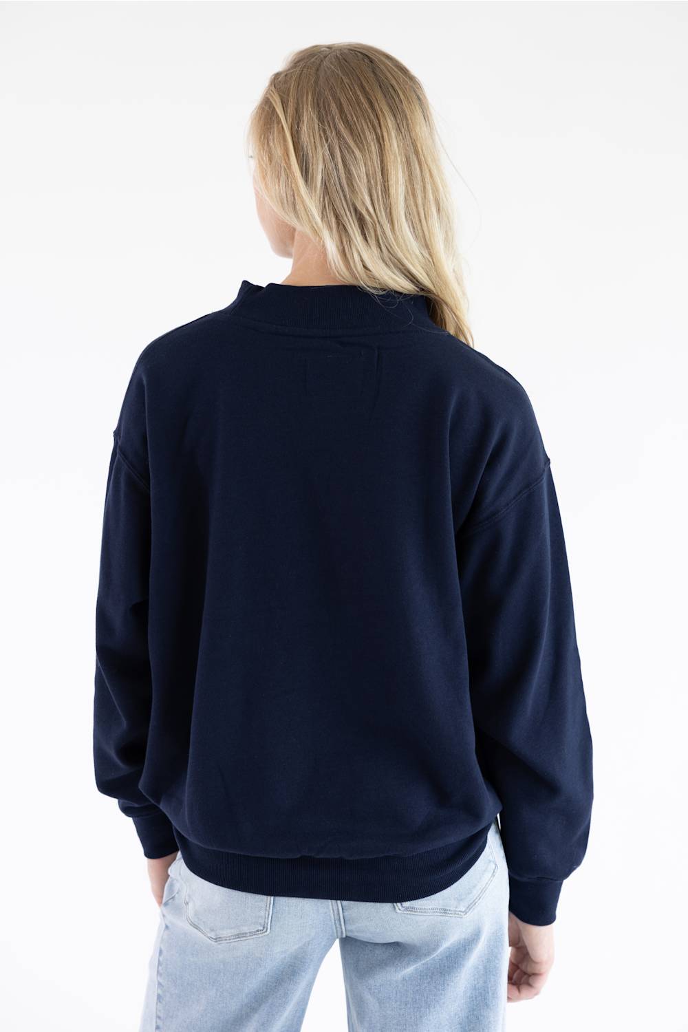 A woman viewed from behind wearing a dark navy blue crewneck sweatshirt and light wash jeans against a plain white background.