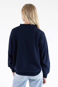A woman viewed from behind wearing a dark navy blue crewneck sweatshirt and light wash jeans against a plain white background.