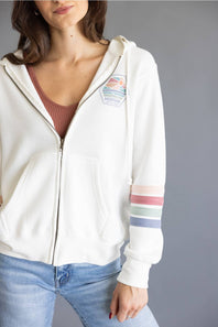 A woman wearing a white zip-up hoodie with a colorful shield patch on the left chest and pastel striped details on the right sleeve, paired with a brown ribbed top and blue jeans against a gray studio background.