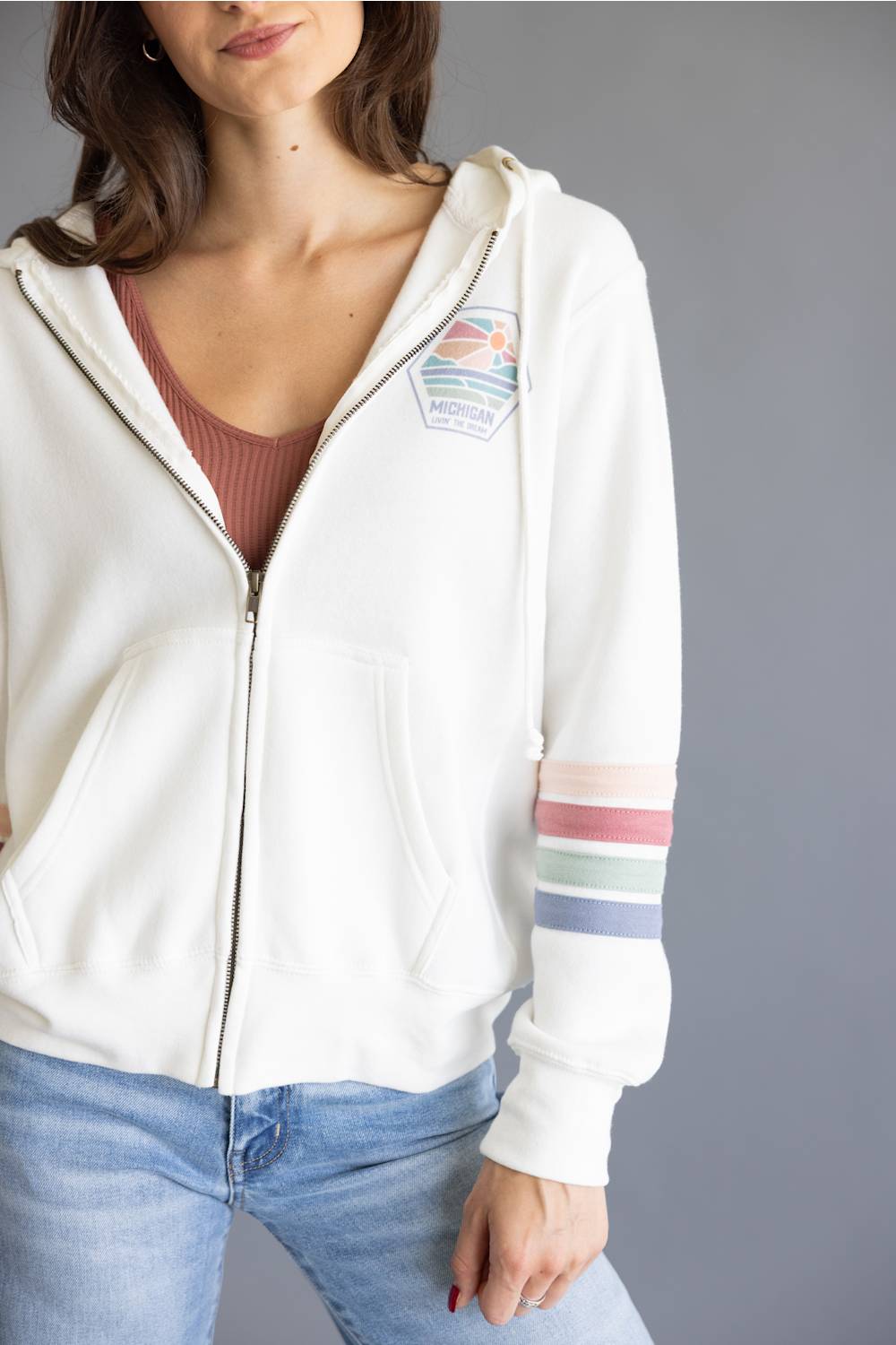 A woman wearing a white zip-up hoodie with a colorful shield patch on the left chest and pastel striped details on the right sleeve, paired with a brown ribbed top and blue jeans against a gray studio background.