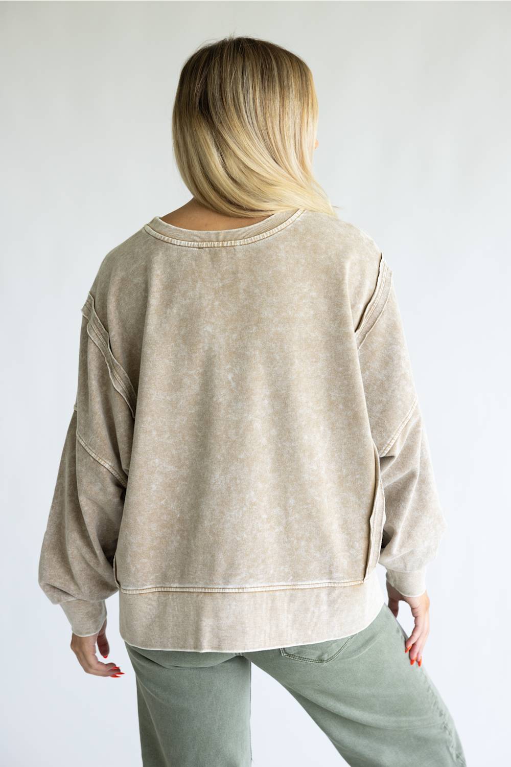A woman viewed from behind wearing a loose, oversized long-sleeve sweatshirt in a light beige mineral-wash fabric, paired with sage green pants, against a plain light gray background.