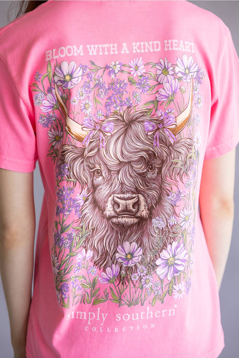 A close-up of a pink short-sleeve t-shirt featuring a large graphic print of a highland cow surrounded by purple flowers, with the text 'BLOOM WITH A KIND HEART' and 'simply southern COLLECTION' on the back.