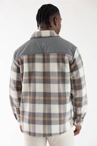 A man viewed from the back wearing a long-sleeve plaid flannel shirt with a solid gray yoke panel on the upper back and shoulders, paired with off-white pants, against a plain white studio background.