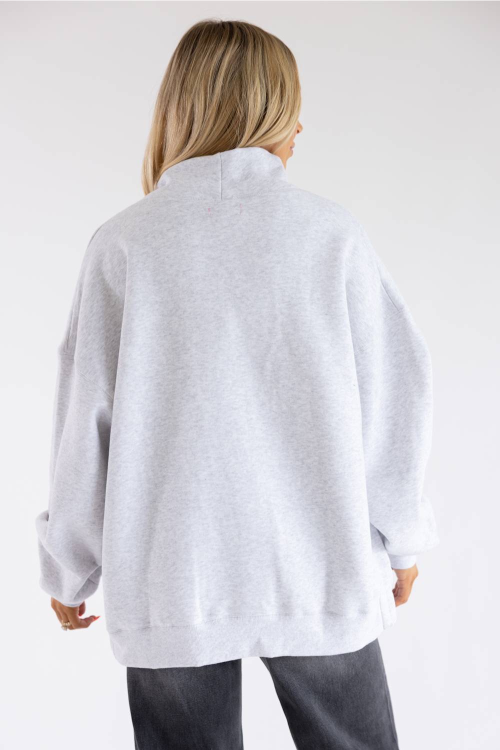 A woman viewed from behind wearing an oversized light gray heather sweatshirt and dark gray jeans against a plain white studio background.
