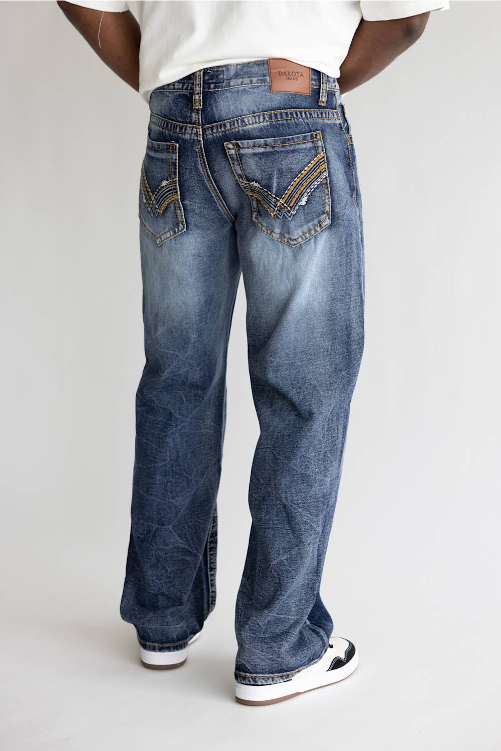 A person shown from the back wearing blue denim jeans with decorative gold embroidery on the back pockets, a white t-shirt, and white sneakers, photographed against a light gray studio background.