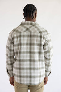 A man wearing a long-sleeve button-up plaid overshirt in beige and olive green, layered over a dark green t-shirt and paired with khaki pants, against a plain white studio background.