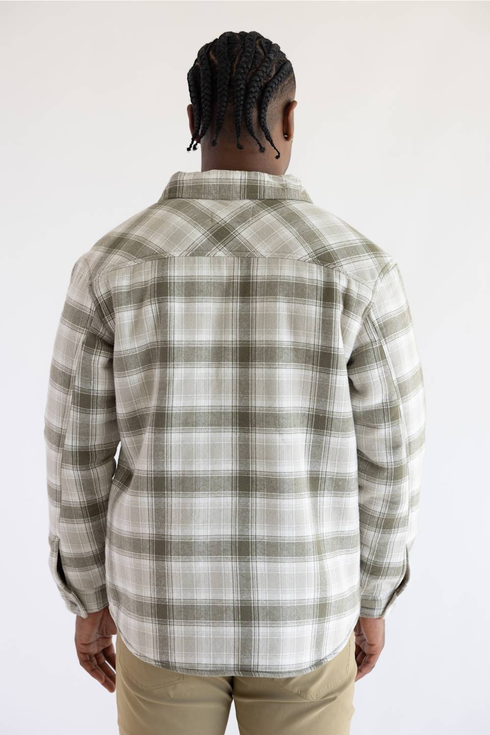 A man wearing a long-sleeve button-up plaid overshirt in beige and olive green, layered over a dark green t-shirt and paired with khaki pants, against a plain white studio background.