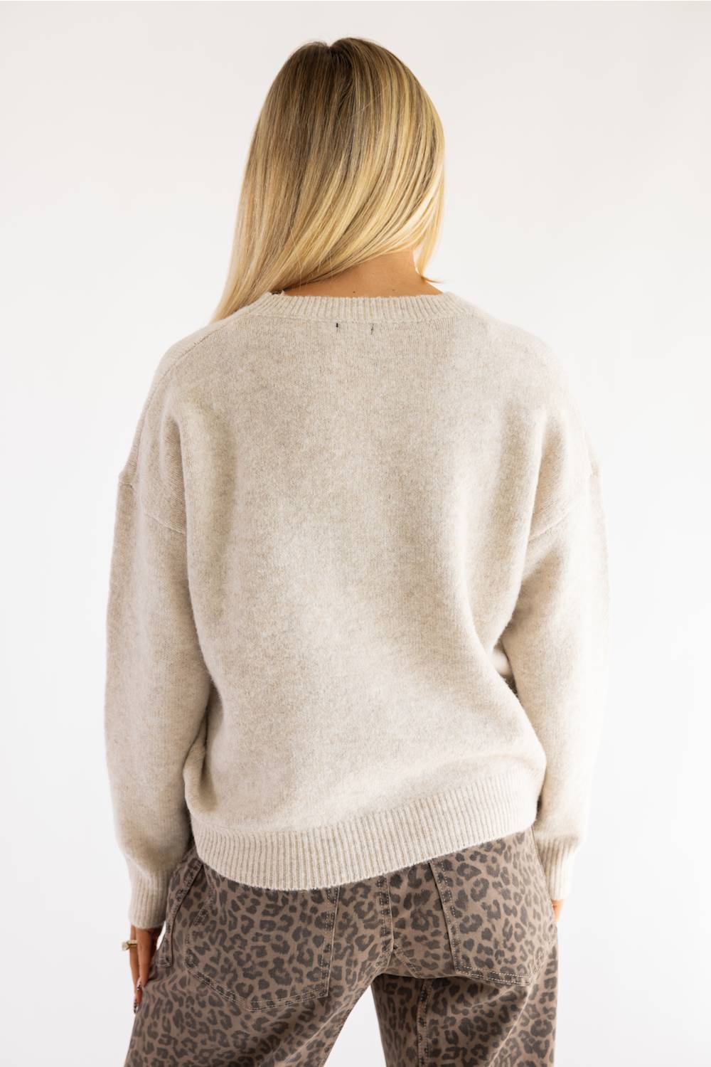 A woman viewed from behind wearing a light beige knit sweater and leopard-print pants against a plain white background.
