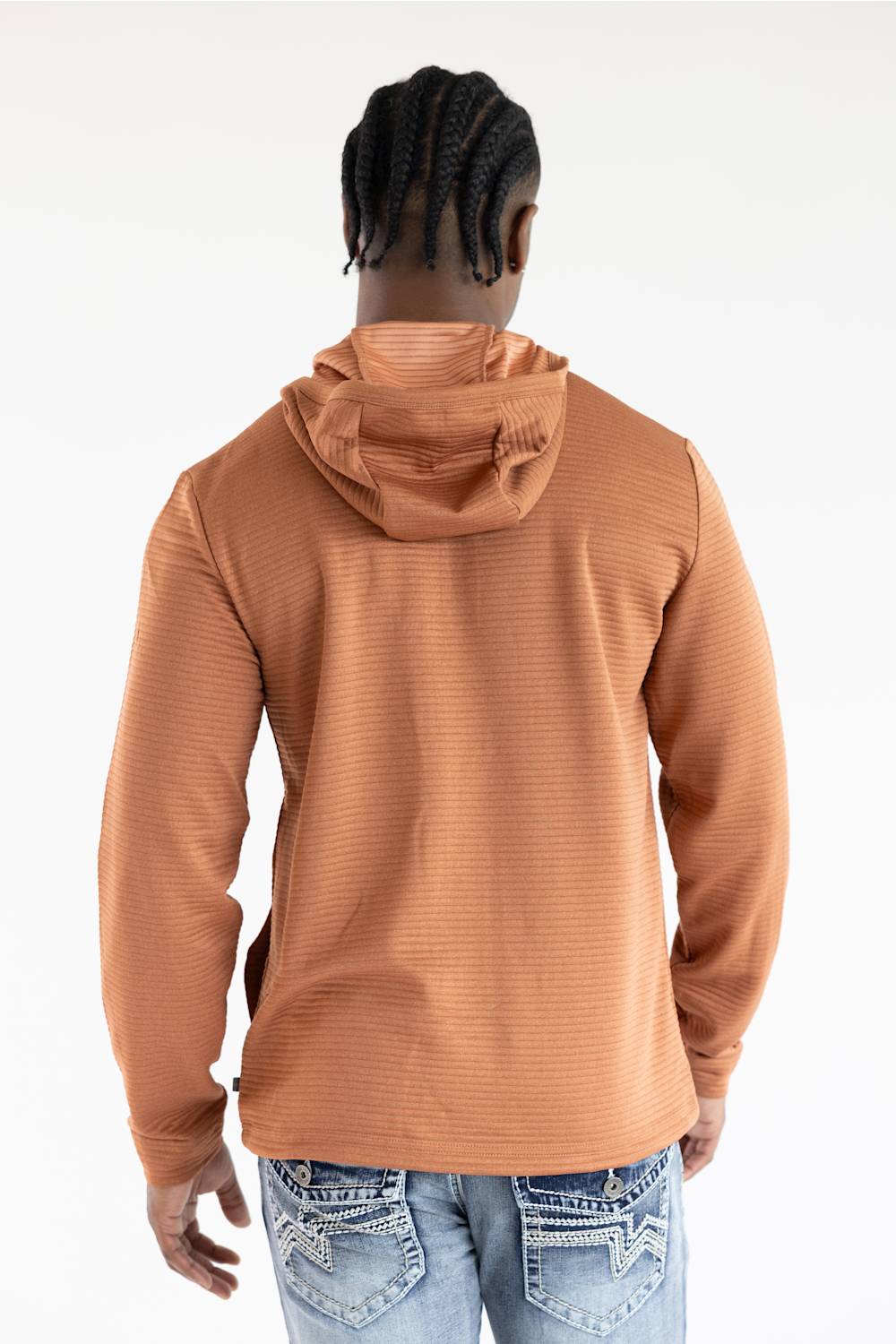 A man viewed from behind wearing a textured, ribbed long-sleeve hooded shirt in a warm terracotta or rust color, paired with light blue jeans, against a plain white studio background.