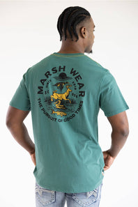 A man wearing a teal green t-shirt with a large back graphic that reads 'MARSH WEAR' and features a yellow deer and black text, shown from behind against a white background.
