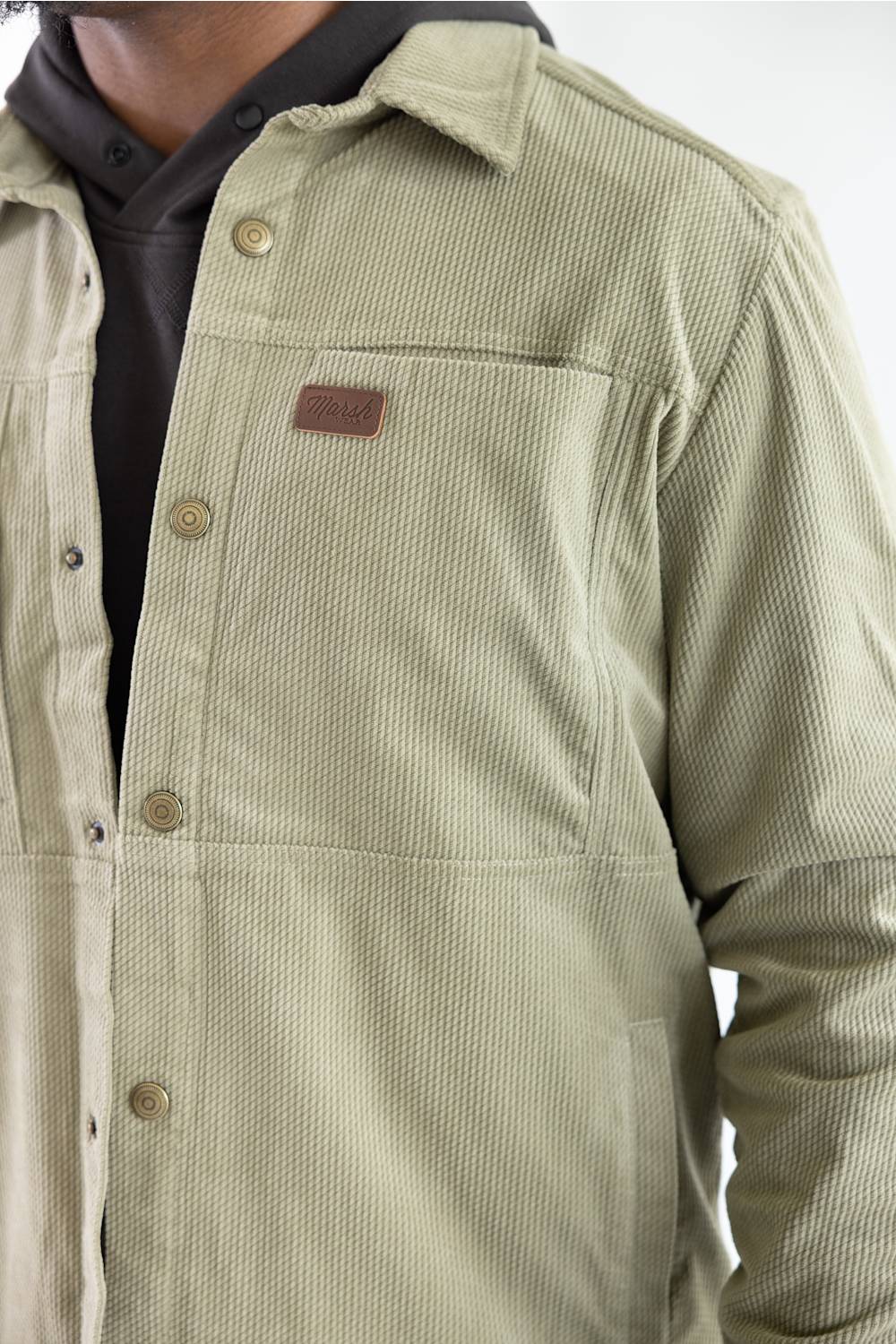 Close-up shot of the Marsh Wear Delano Cord Shacket in Sage Brush green corduroy, showcasing the chest pocket with an embossed leather brand patch, antique brass snap buttons, collar detail, and textured diamond check pattern.
