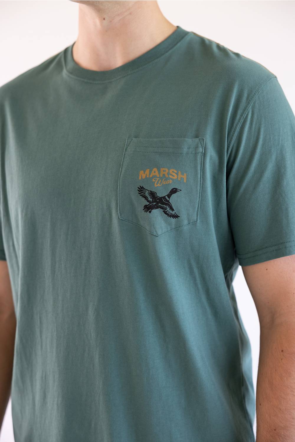 A man wearing a muted teal-green pocket t-shirt with a black embroidered duck and gold 'MARSH Lake' text on the chest pocket.