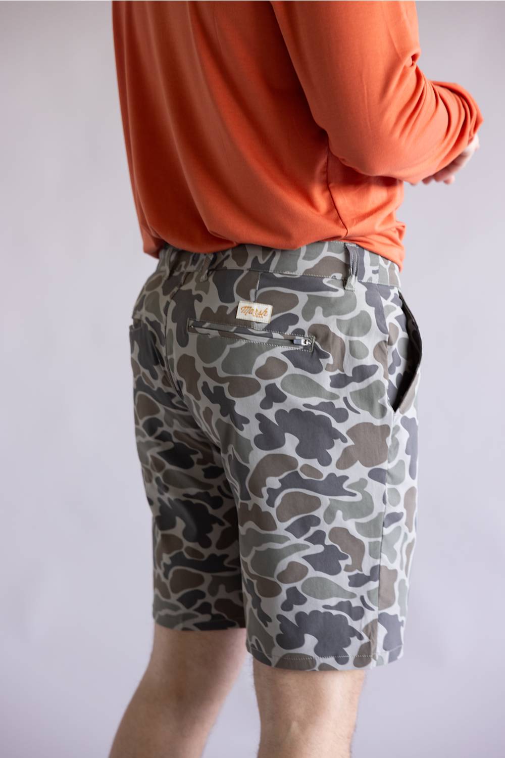 Marsh Wear Prime Hagood 8” Shorts for Men in Green Mallard Camo; Person wearing camouflage shorts and an orange long-sleeve shirt on a light gray background
