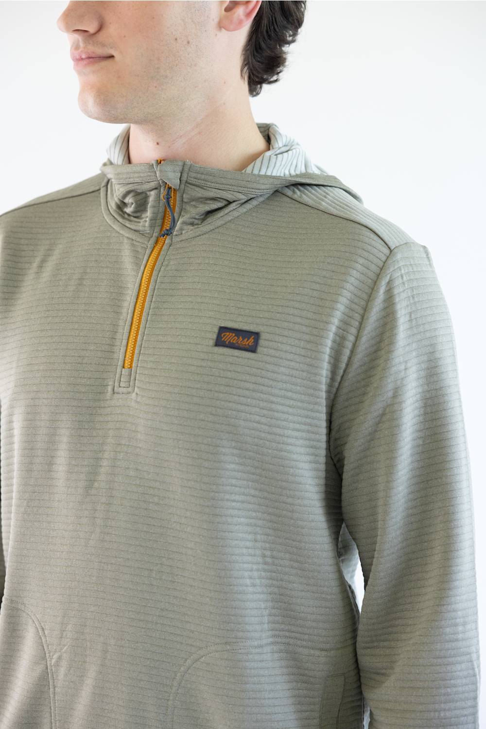 A man wearing a Marshwear hooded quarter-zip pullover in a textured sage green fabric with a small orange zipper and a small Marshwear  logo patch on the chest, paired with blue jeans against a plain white background.