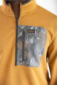 Close-up detail shot showing the textured ribbed fabric of the Marsh Wear Sullivan Quarter Zip Pullover in Buckthorn Brown, featuring a grey zip chest pocket with a leaf camouflage pattern, orange "Marsh Wear" script logo patch, and front quarter-zip closure.