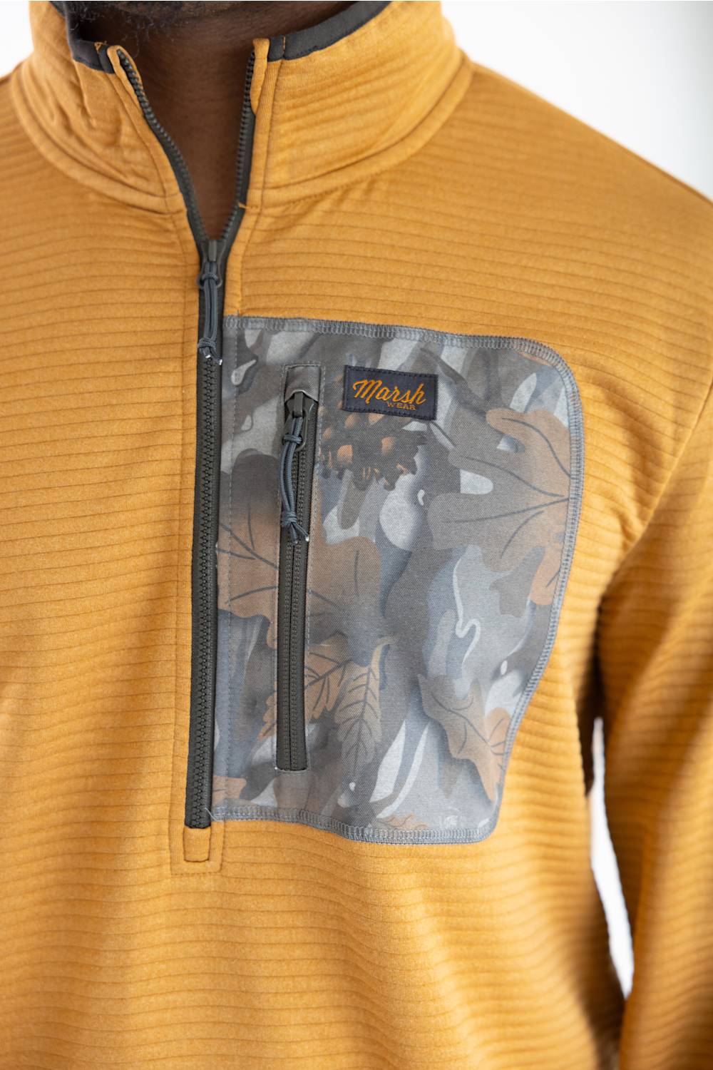 Close-up detail shot showing the textured ribbed fabric of the Marsh Wear Sullivan Quarter Zip Pullover in Buckthorn Brown, featuring a grey zip chest pocket with a leaf camouflage pattern, orange "Marsh Wear" script logo patch, and front quarter-zip closure.