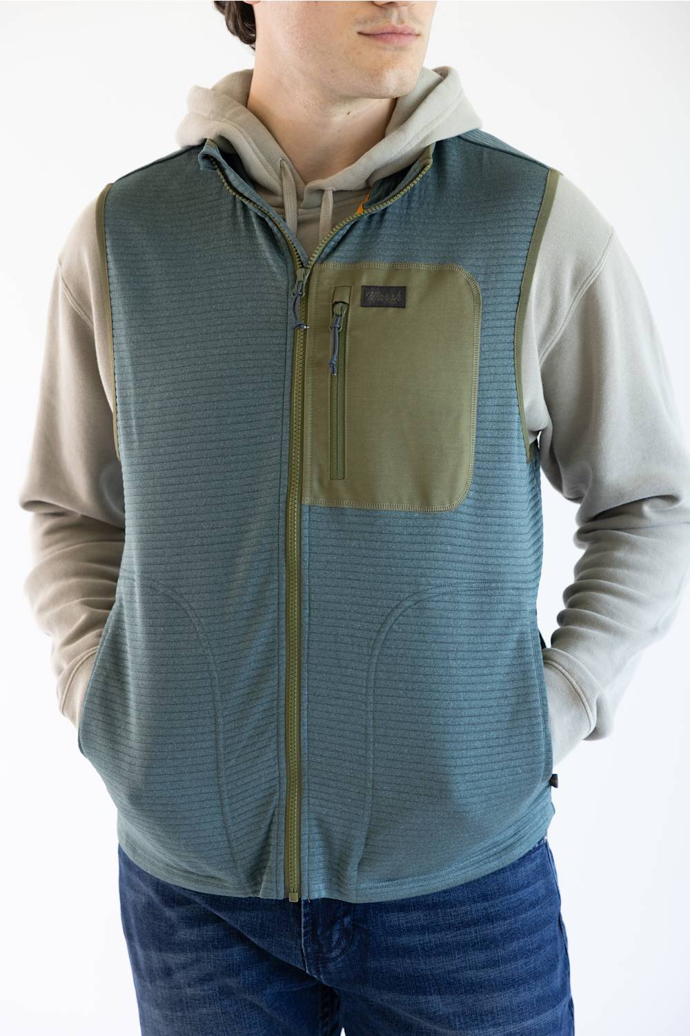 A man wearing a sleeveless zip-front vest over a light beige hoodie and blue jeans. The vest is slate blue with olive green trim and a matching olive chest pocket, shown against a plain white background.