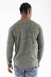 Back view of a model wearing the Marsh Wear Surfside Waffle Knit Shirt in Fossil Foxhole Camo green, showcasing the long-sleeve waffle-textured fabric and raglan shoulder seam detailing, paired with light-wash blue jeans featuring decorative back pocket stitching