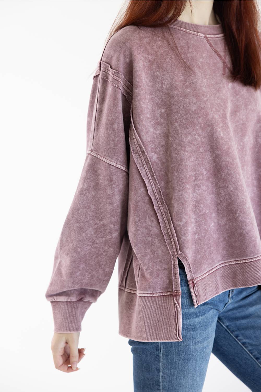 A person wearing a faded dusty rose long-sleeve sweatshirt with diagonal seam details and side slits, paired with blue jeans, against a plain white background.