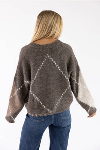 Back view of a woman with long blonde hair wearing a brown Miracle argyle V-neck sweater featuring a soft fuzzy knit fabric and an all-over diamond pattern, paired with dark denim jeans.