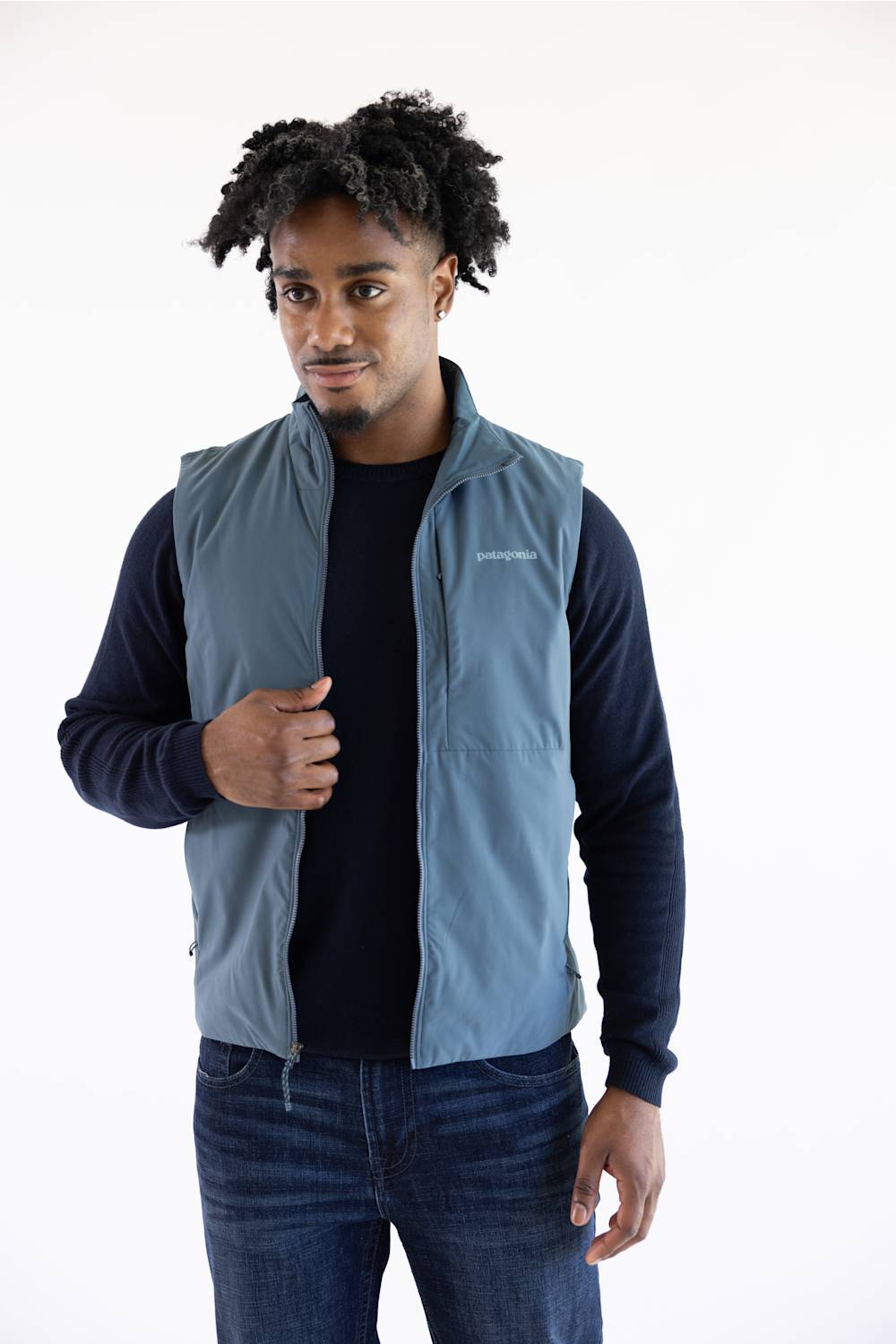 A man wearing a slate blue zip-up vest over a dark navy long-sleeve shirt and blue jeans, standing against a plain white studio background.