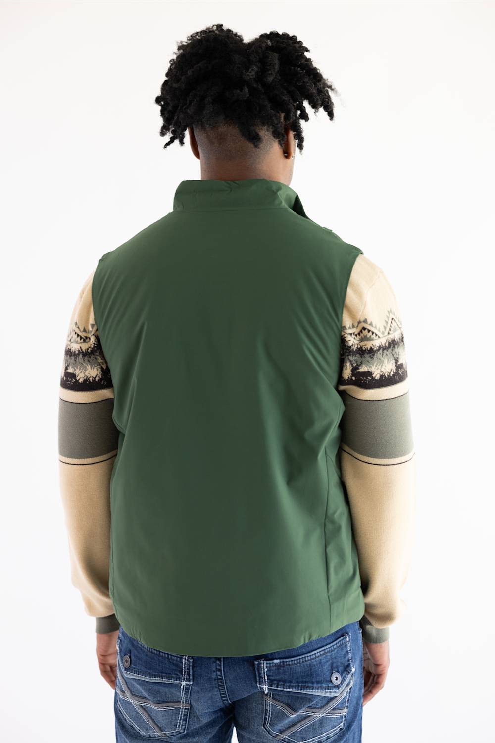 A person shown from the back wearing an olive green sleeveless vest layered over a beige and gray patterned long-sleeve shirt, paired with blue jeans, against a plain white background.