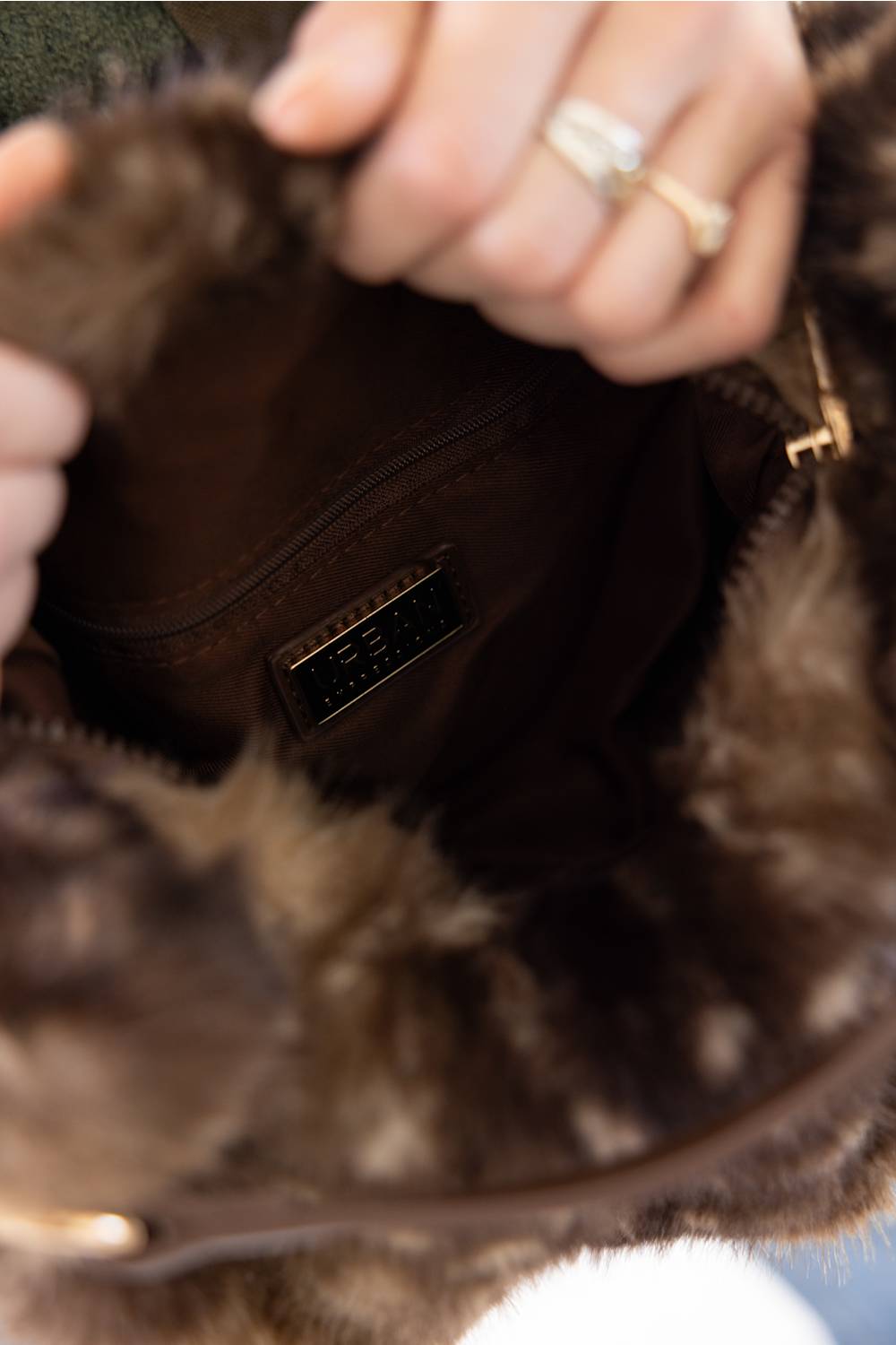 Close-up of a brown fur or faux-fur handbag interior with a person's hands reaching inside, showing a dark brown lining and a small black brand plaque.