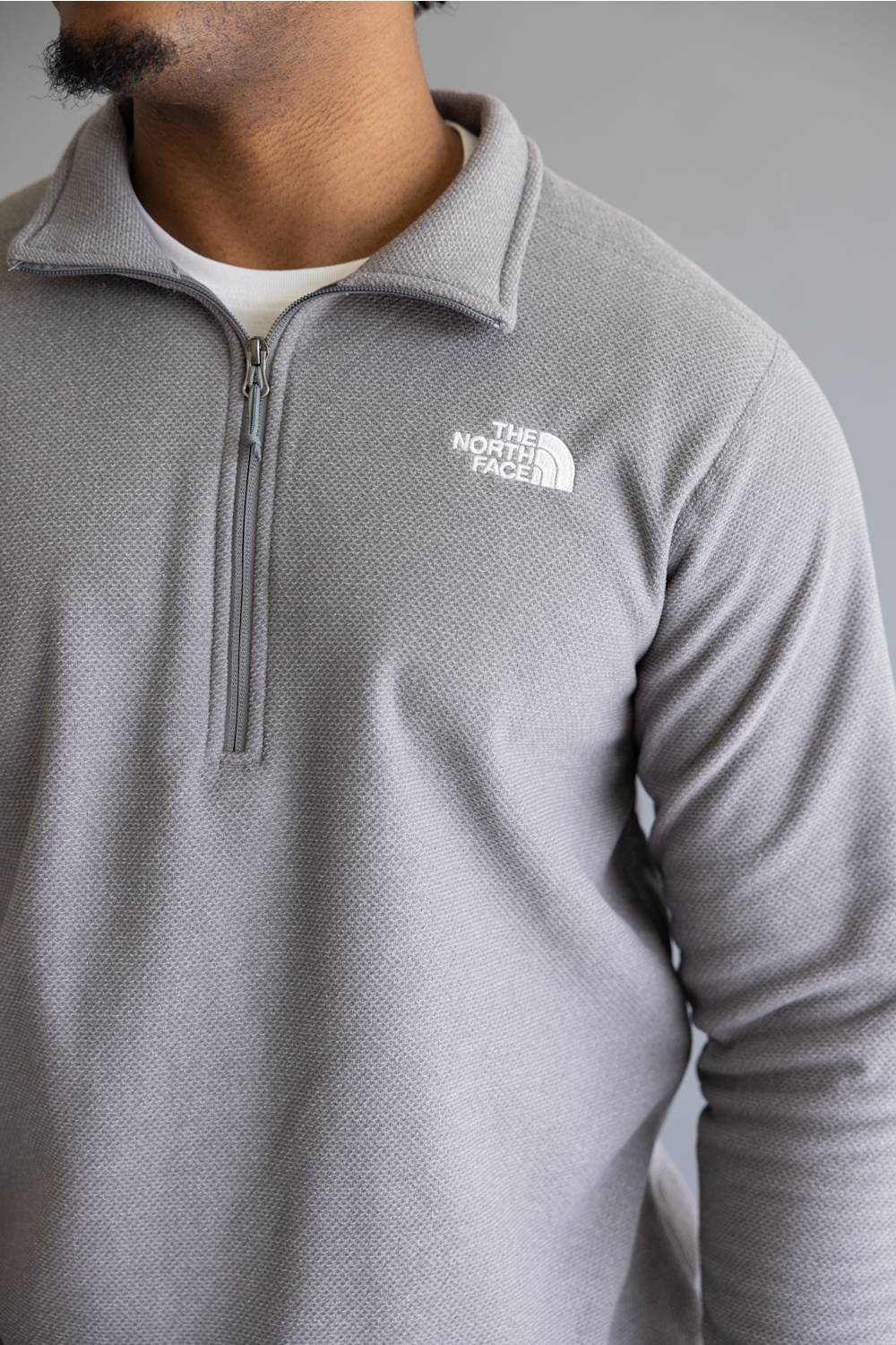 A close-up of a man wearing a light gray heathered quarter-zip pullover from The North Face, featuring a white embroidered logo on the left chest.