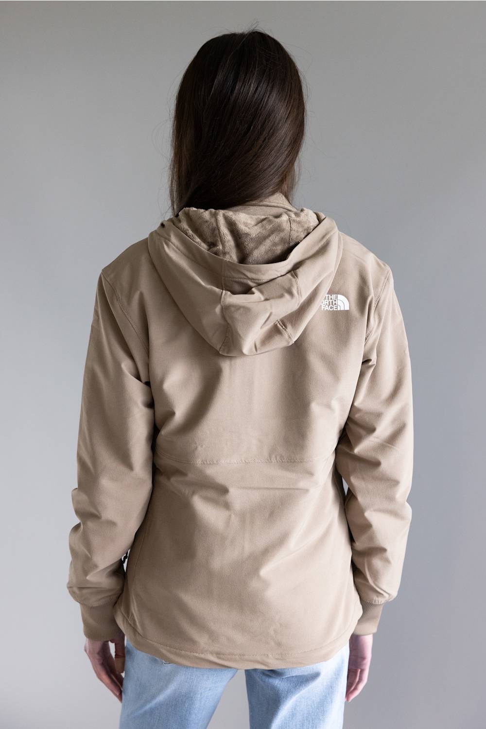 A person shown from the back wearing a beige hooded jacket with a small white The North Face logo on the upper back, paired with light blue jeans, against a plain gray studio background.