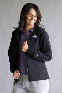A woman wearing a black The North Face zip-up jacket over a purple collared shirt and gray sweatpants, standing against a neutral gray studio background.