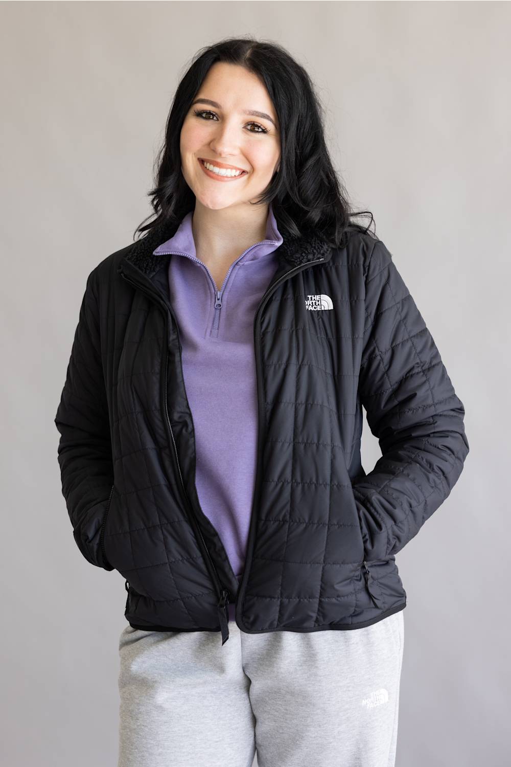 A woman smiling and wearing a black quilted puffer jacket by The North Face over a purple quarter-zip pullover and light gray sweatpants, set against a plain light gray studio background.