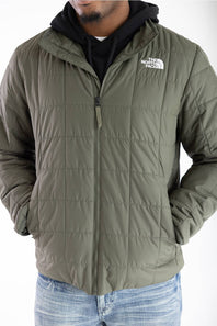 An olive green quilted puffer jacket by The North Face, worn by a model over a black hoodie and blue jeans against a white background.