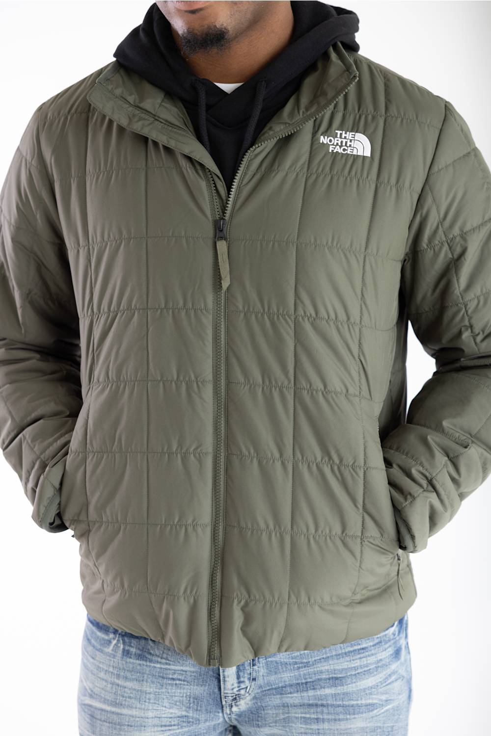 An olive green quilted puffer jacket by The North Face, worn by a model over a black hoodie and blue jeans against a white background.